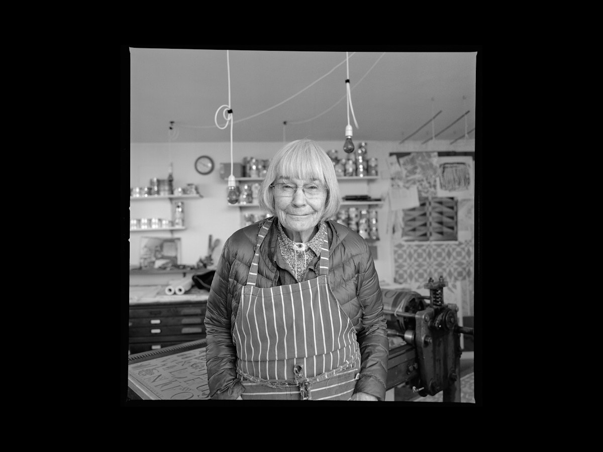 An older lady standing with a striped apron on. She has grey hair and stares at the camera.