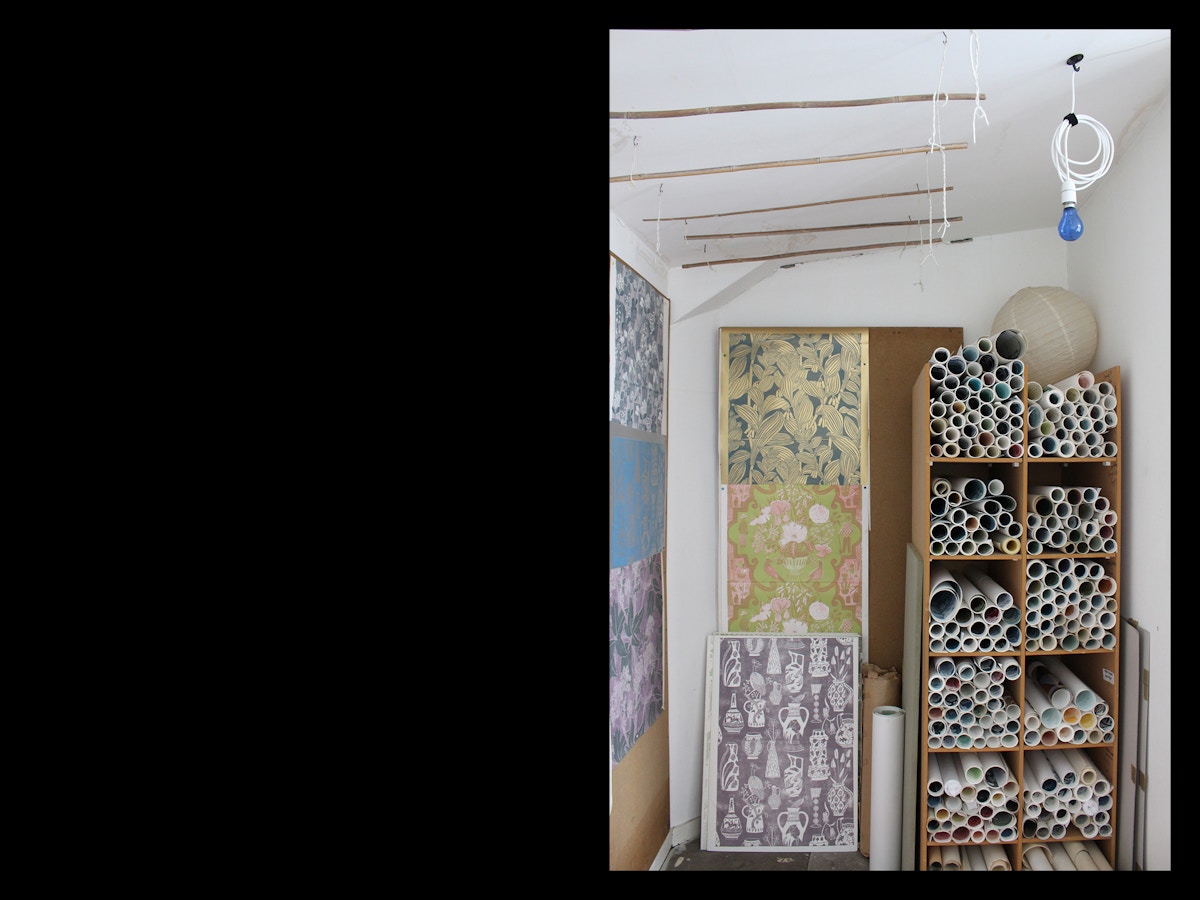 Rolls of wallpaper in a wooden shelving system. There are other pieces of wallpaper stuck on a wall beside it.