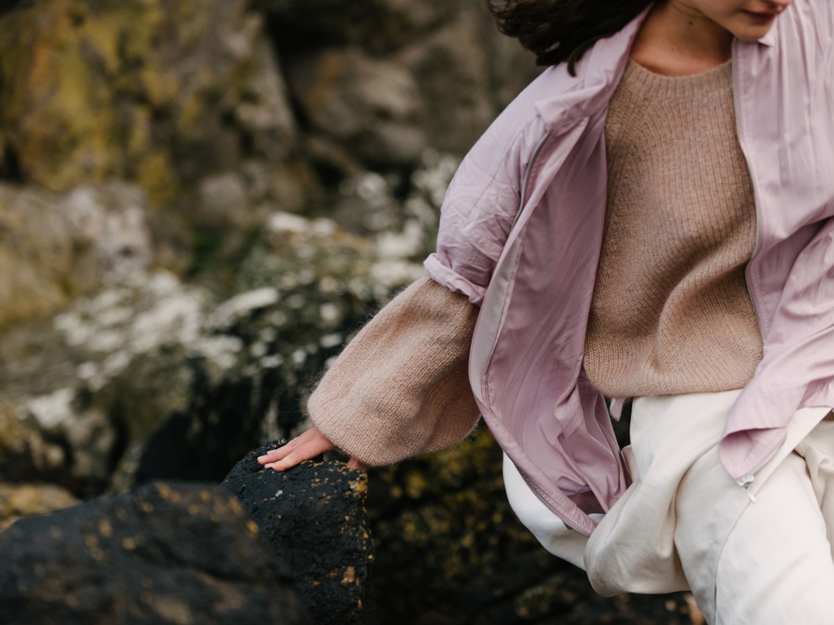 A woman in a beige jumper and pink jacket leaning against some rocks.