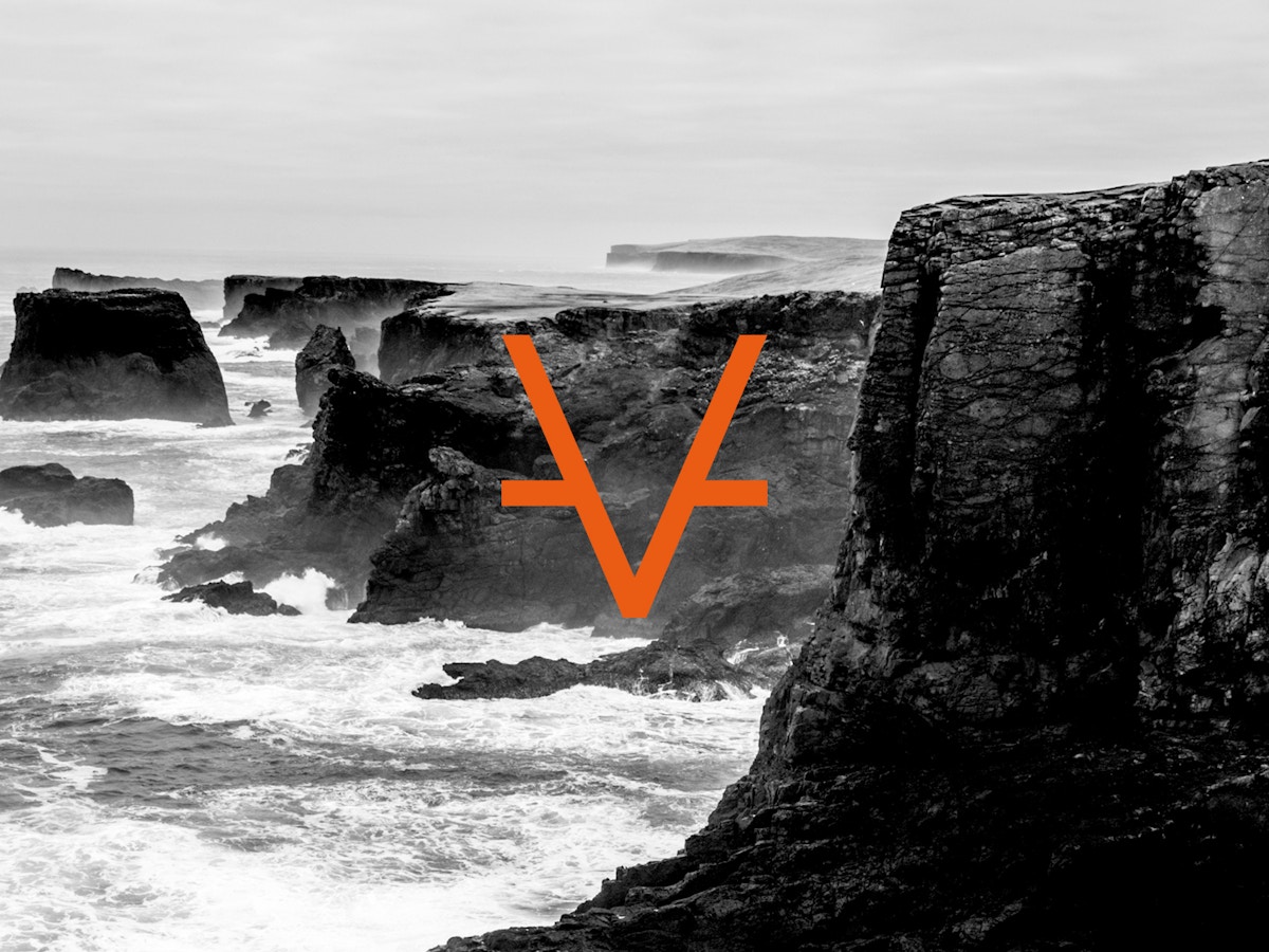 Cliff face with crashing waves in black and white. There is an orange V on top.