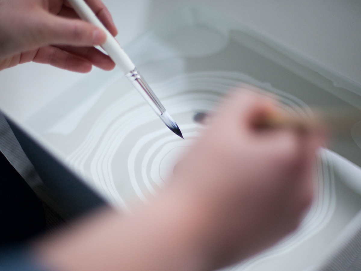 Two hands holding paintbrushes above a swirled pattern made in water.