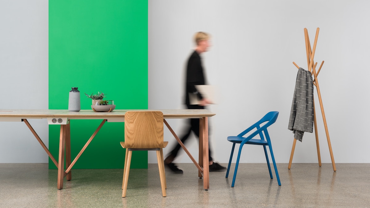A table and chair set up in front of a green panel. There is a blurred person walking out of shot in the background