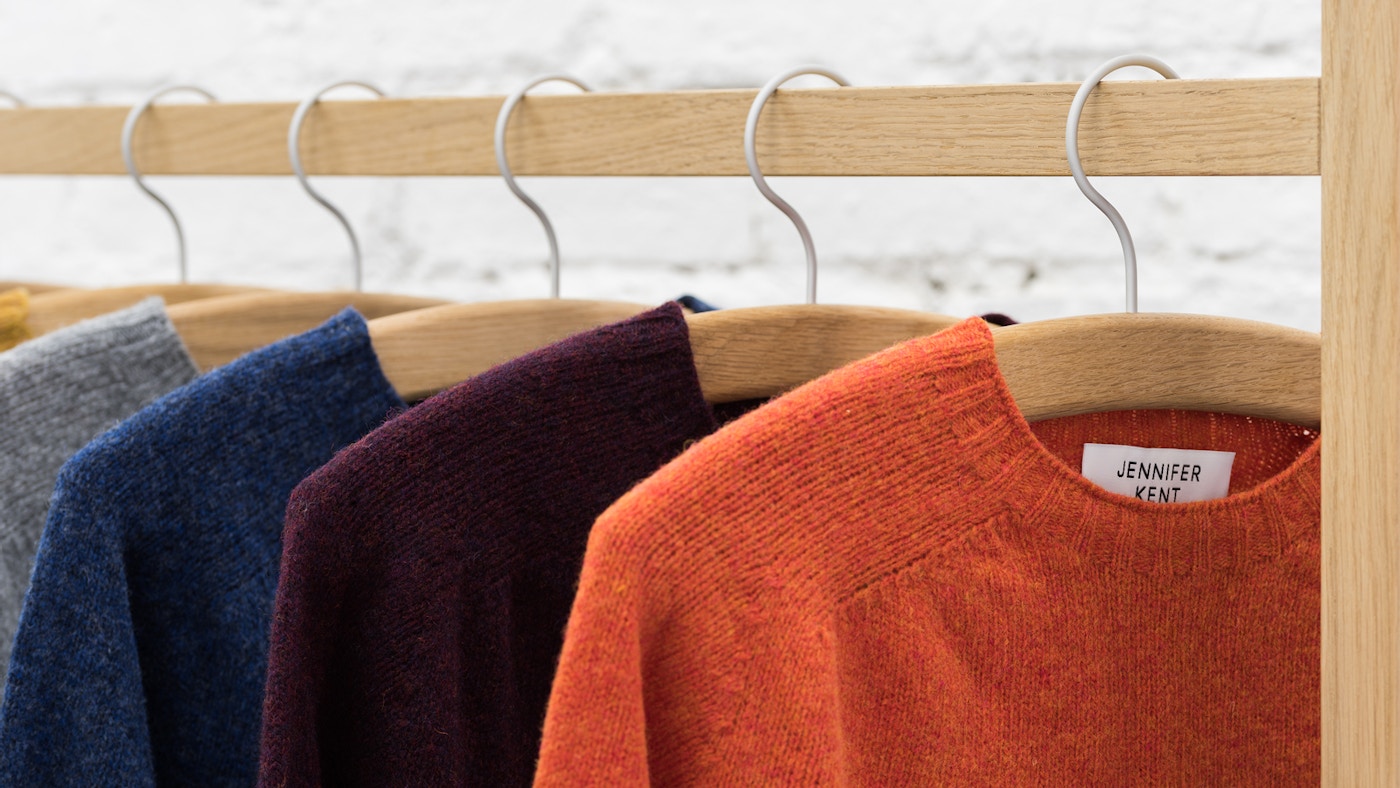 Four jumpers sitting on wooden hangers on a wooden rail. The jumpers are orange, purple, blue and grey.
