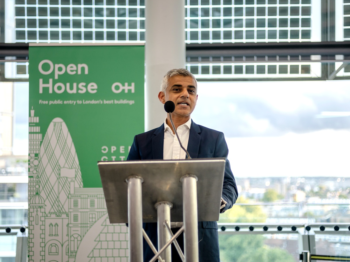 A man stands at a podium in front of a banner that reads "Open House" and has an illustration of the London skyline on it.