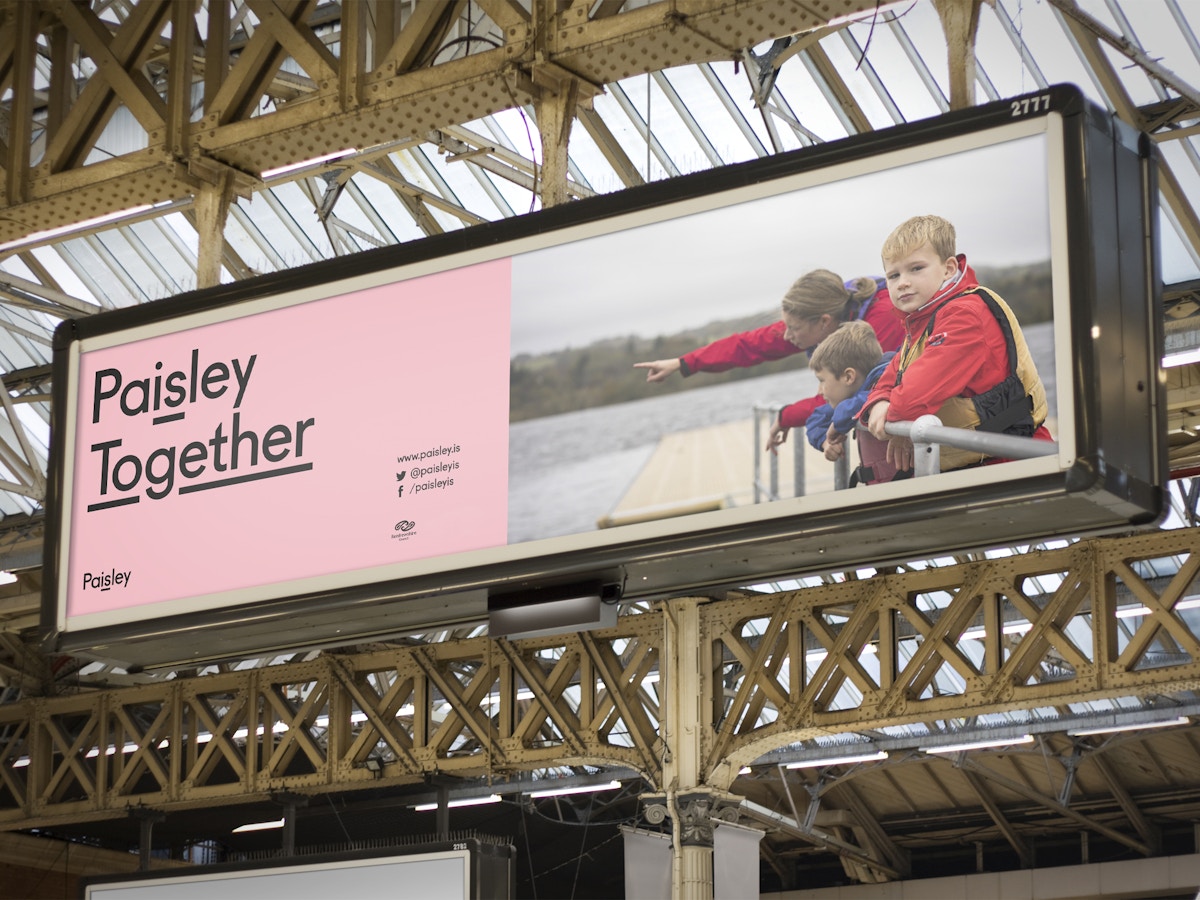 A billboard advertisement in a train station. The text reads "Paisley Together" set in black type on a pink background.
