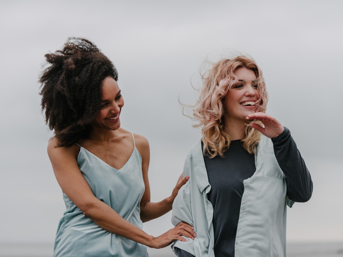 Two women laughing. Their hair appears to be blowing in the wind. The woman on the left is holding onto the others arm.