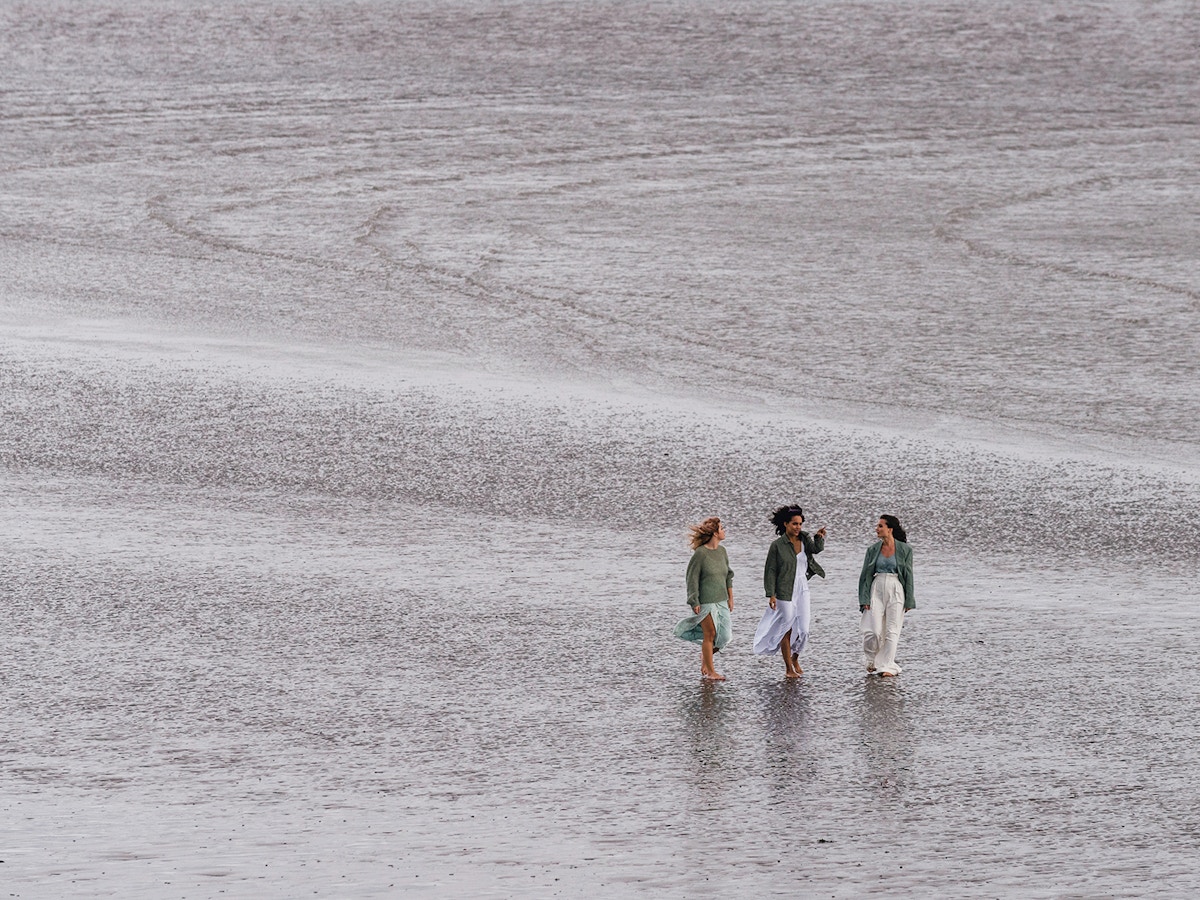 Three women walking beside each other along a beach. There is only sand visible behind them.