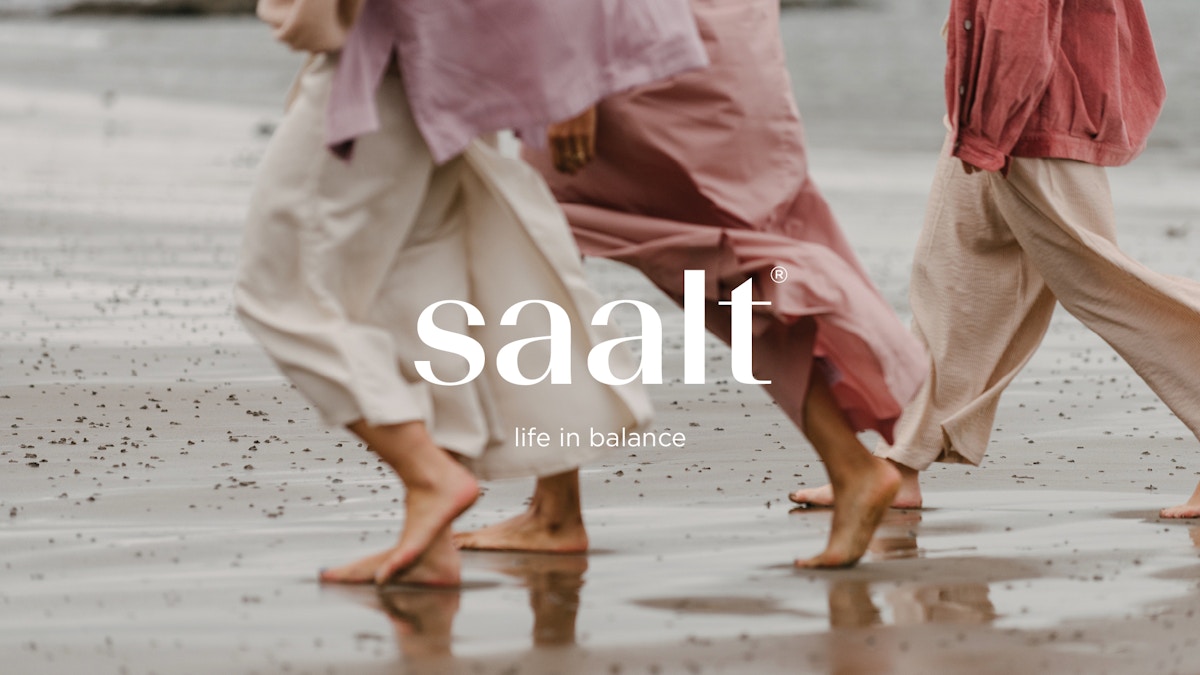 Text that reads "Saalt. Life in balance" over a photograph of three women walking barefoot along a beach. They are all wearing pinky red shades of clothes.