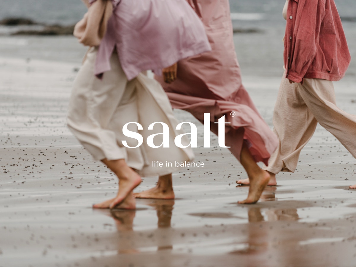 Text that reads "Saalt. Life in balance" over a photograph of three women walking barefoot along a beach. They are all wearing pinky red shades of clothes.