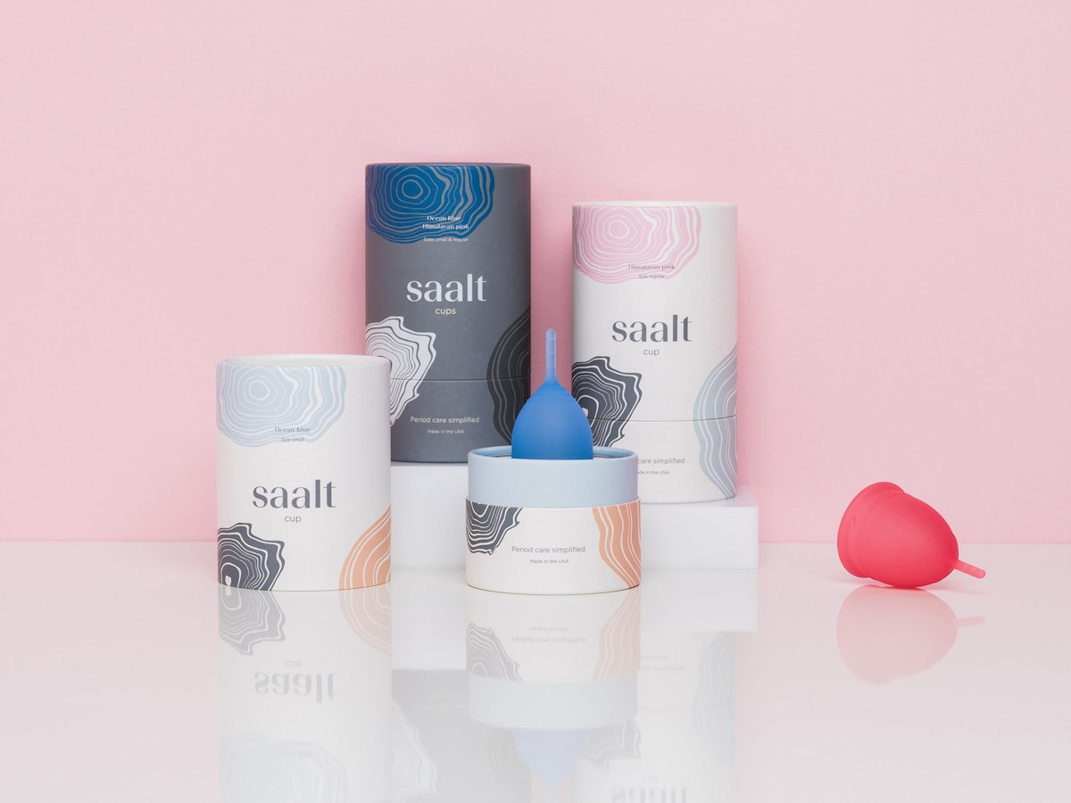 Three Saalt packaging tubes in front of a pink background. There is a blue menstrual cup sitting centrally and a pink one to the right.