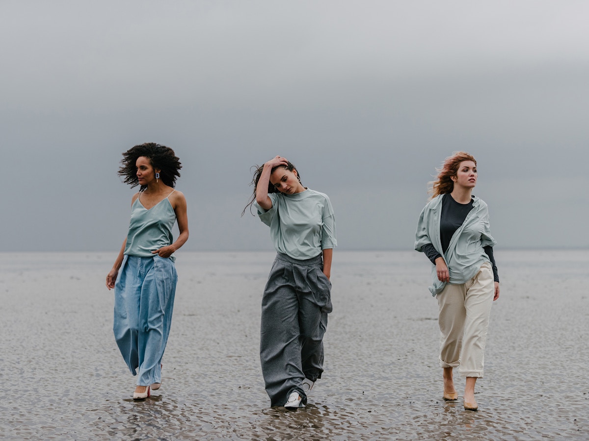 Three women walking along a beach towards the camera. The woman in the middle has her right hand on her head. They are all wearing shades of blue.
