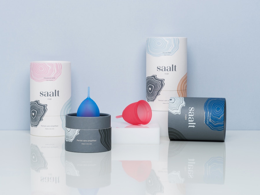 Three Saalt packaging tubes sitting in front of a blue background. There is a blue and a red menstrual cup sitting in the centre.