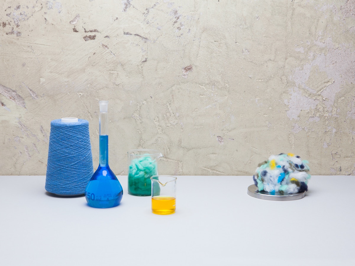 A loom, bottle with blue liquid, glass with yellow liquid and a ball of multicoloured fabric sitting on a white tabletop.