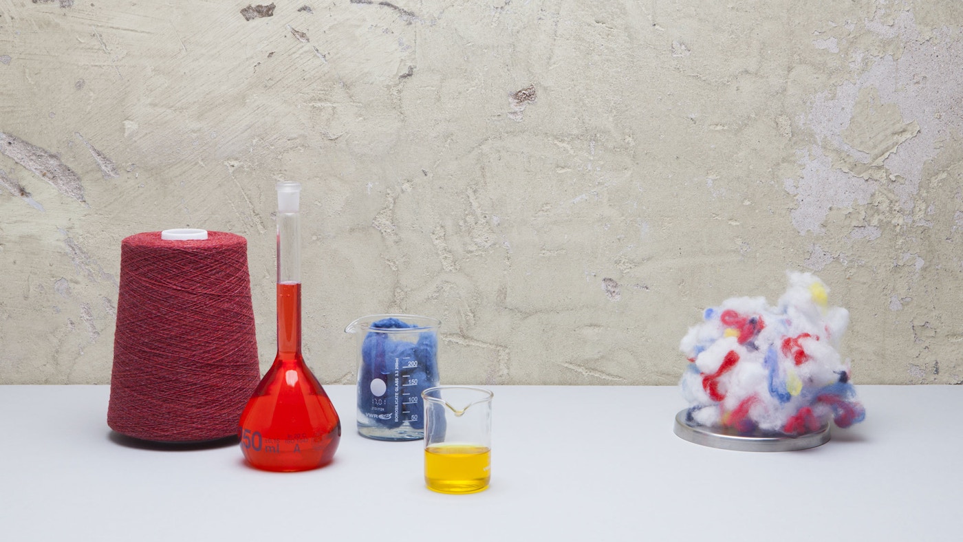 A loom, bottle with red liquid, glass with yellow liquid and a ball of multicoloured fabric sitting on a white tabletop.